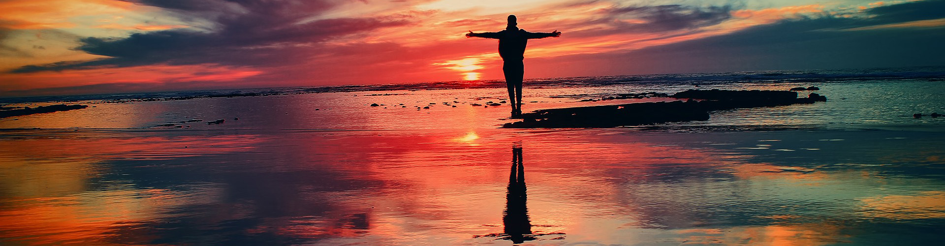 Man standing with his arms open on a beach with a red sunset in the background